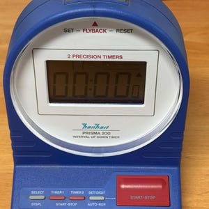 May include: Blue Hanheart Prisma 200 interval up-down timer with a digital display showing 00:00. Features buttons for set, flyback, reset, start-stop, and timer selection. The timer is on a wooden surface.