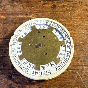 May include: A white circular calendar disc with black text and numbers. The disc has a gold-colored center with a circular pattern and three holes. The text on the disc reads: "SUNDAY MONDAY TUESDAY WEDNESDAY THURSDAY FRIDAY SATURDAY". The numbers on the disc range from 1 to 31.