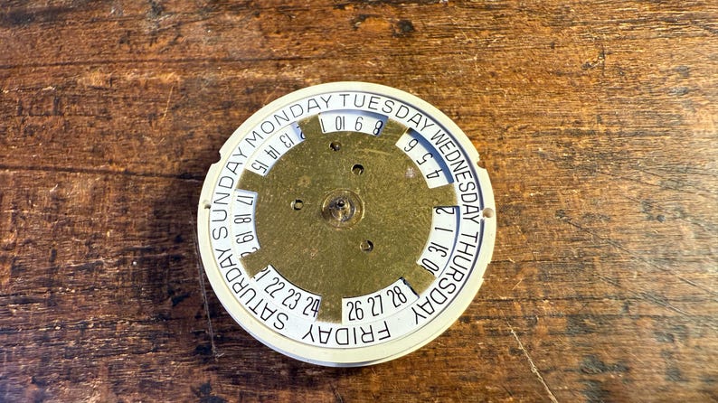 May include: A white circular calendar disc with black text and numbers. The disc has a gold-colored center with a circular pattern and three holes. The text on the disc reads: "SUNDAY MONDAY TUESDAY WEDNESDAY THURSDAY FRIDAY SATURDAY". The numbers on the disc range from 1 to 31.
