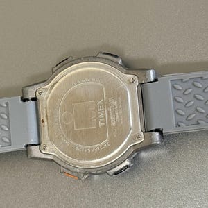 K&ouml;nnte beinhalten: Eine silberfarbene Timex Digitaluhr mit einem grauen Kautschukarmband. Das Armband hat ein strukturiertes, rautenf&ouml;rmiges Muster. Das Zifferblatt ist nicht sichtbar, aber die R&uuml;ckseite der Uhr ist zu sehen, mit dem Timex-Logo und anderem Text.