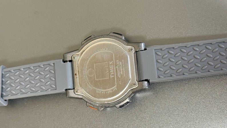 K&ouml;nnte beinhalten: Eine silberfarbene Timex Digitaluhr mit einem grauen Kautschukarmband. Das Armband hat ein strukturiertes, rautenf&ouml;rmiges Muster. Das Zifferblatt ist nicht sichtbar, aber die R&uuml;ckseite der Uhr ist zu sehen, mit dem Timex-Logo und anderem Text.