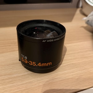 以下が含まれることがあります： 「1:1.8 5.9-35.4mm」と「AF WIDE-ANGLE ZOOM」というオレンジ色の文字が書かれた黒いカメラレンズです。