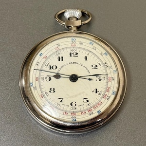 Könnte beinhalten: Eine silberne Vintage-Taschenuhr mit weißem Zifferblatt und schwarzen Zeigern. Das Zifferblatt hat schwarze Ziffern, rote und blaue Markierungen und die Worte "CHRONOGRAPHE DE SPORT". Die Uhr hat eine Krone oben.