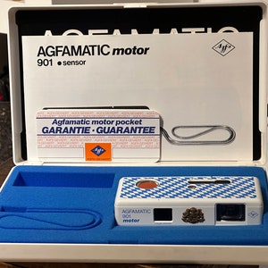 Può includere: Una macchina fotografica Agfamatic 901 motor bianca e blu nella sua scatola originale. La macchina fotografica ha un design a scacchi blu e bianco e la scatola ha un interno blu.