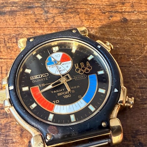 Könnte beinhalten: Vintage Seiko Quartz-Uhr mit schwarzem Zifferblatt, goldenen Akzenten und einem schwarzen und goldenen Armband. Die Uhr verfügt über ein USA-Olympia-Logo und eine Yacht-Timer-Funktion. Das Zifferblatt hat rote, weiße und blaue Akzente.