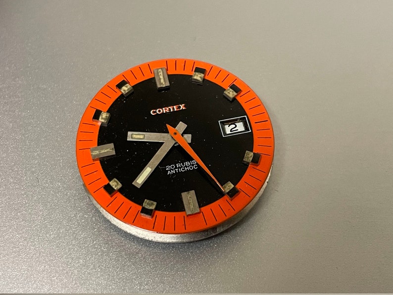 K&ouml;nnte beinhalten: Ein schwarzes Zifferblatt mit einem orangefarbenen Au&szlig;enring und wei&szlig;en Markierungen. Das Zifferblatt der Uhr zeigt "CORTEX 20 RUBIS ANTICHOC" und hat ein kleines Datumsfenster, das die Zahl "2" anzeigt.