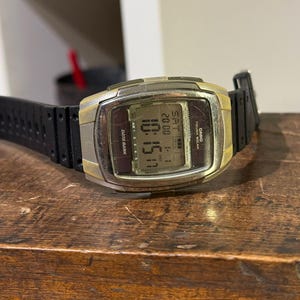 Può includere: Un orologio da polso digitale con cinturino nero e quadrante dorato e argentato. L'orologio mostra l'ora, la data e il giorno della settimana. L'orologio è appoggiato su una superficie di legno.