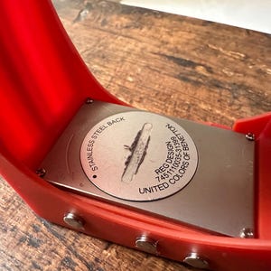 Op de afbeelding: De achterkant van een rood horloge met een zilveren roestvrijstalen achterkant. De achterkant is gegraveerd met "Stainless Steel Back", "Reg. Design 745110035-31399" en "United Colors of Benetton".