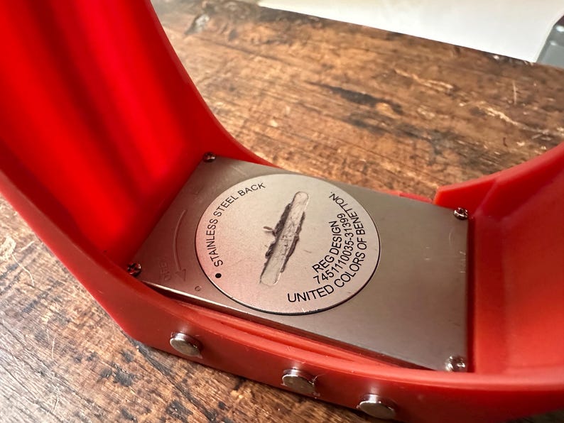 Op de afbeelding: De achterkant van een rood horloge met een zilveren roestvrijstalen achterkant. De achterkant is gegraveerd met "Stainless Steel Back", "Reg. Design 745110035-31399" en "United Colors of Benetton".