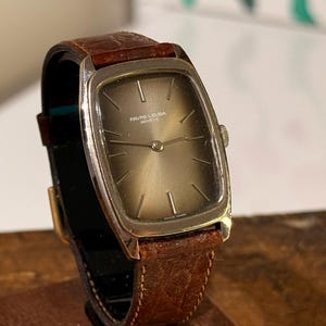 May include: A vintage wristwatch with a rectangular silver-toned face and a brown leather strap. The watch face has a light brown hue with silver hands and markers. The strap is a dark brown with a textured pattern.