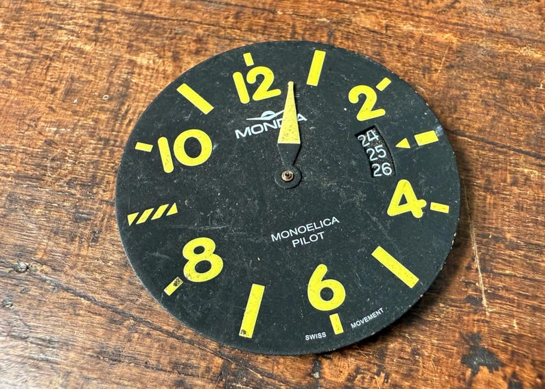 May include: A black watch face with yellow numbers and markings. The watch face reads "10, 8, 12, 2, 4, 6" and "MONOELICA PILOT" with "SWISS MOVEMENT".