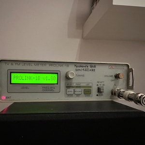 以下が含まれることがあります： 「PROLINK-1B v1.80」と表示された緑色のデジタルディスプレイを持つ、白いProlink-1B TVおよびFMレベルメーター。メーターには、チューニング、ボリューム、RF減衰用のノブがあります。また、バッテリーインジケーターライトと同軸ケーブルコネクターも備えています。