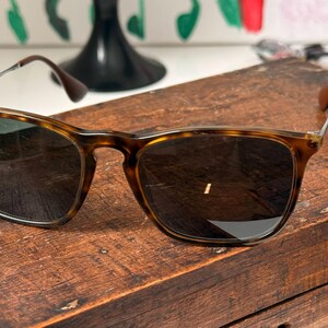 May include: Tortoiseshell pattern sunglasses with dark lenses. The sunglasses are on a wooden surface. The frames have a classic, rectangular shape with a brown and tan pattern. The lenses are dark.