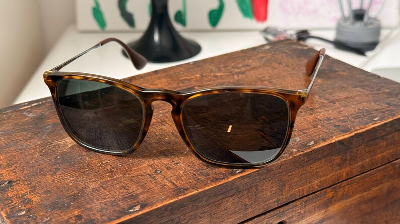 May include: Tortoiseshell pattern sunglasses with dark lenses. The sunglasses are on a wooden surface. The frames have a classic, rectangular shape with a brown and tan pattern. The lenses are dark.