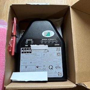 May include: A black KONE4G LTE device with a signal strength indicator, status lights, and a pin connector. The device is in a cardboard box with a white piece of paper and a red cable.