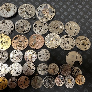 Könnte beinhalten: Eine Sammlung von 35 silbernen und goldenen Uhrwerken, einige mit filigranen Details, in einem Raster auf einer schwarzen Oberfläche angeordnet.