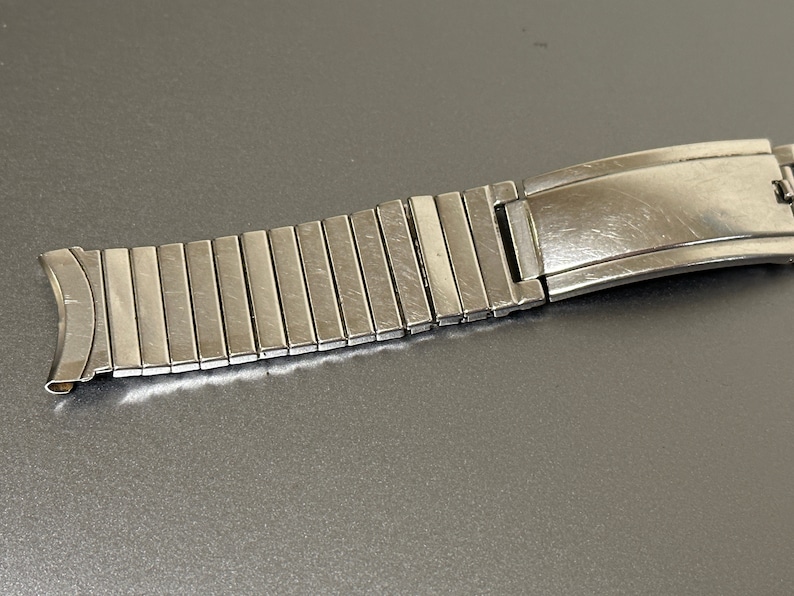 May include: A silver watch band with a clasp. The band is made of many small, linked pieces.