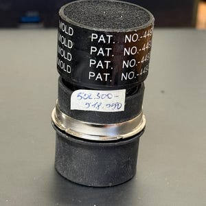 May include: A black cylindrical object with a textured top and a silver band. The object has the word "MOLD" printed on a black band, along with "PAT. NO.-449". A small white label displays the numbers "502.300- 518.250".