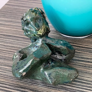 May include: A green ceramic sculpture of a person with a textured surface. The sculpture is sitting on a wooden surface.