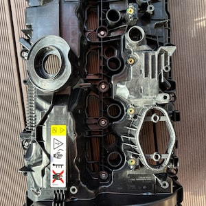 Può includere: Coperchio valvola motore in plastica nera con un'etichetta di avvertenza gialla che recita "WARNING HOT SURFACE" e un'etichetta di avvertenza rossa che recita "DANGER HOT SURFACE".
