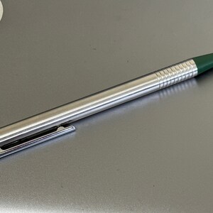 Puede incluir: Un bol&iacute;grafo plateado con una punta verde y una banda verde cerca de la parte inferior. El bol&iacute;grafo tiene un agarre texturizado.