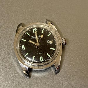 Op de afbeelding: Een vintage polshorloge met een zwarte wijzerplaat en een zilverkleurige kast. De wijzerplaat van het horloge heeft lichtgevende uurmarkeringen en wijzers, met de merknaam "Emcasatic" zichtbaar. Het horloge heeft een datumvenster op 3 uur.