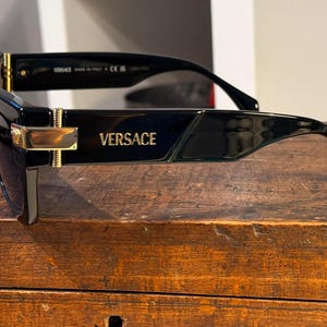 May include: Black Versace sunglasses with a rectangular frame and gold accents. The brand name is visible on the temple. The sunglasses are set on a wooden surface.