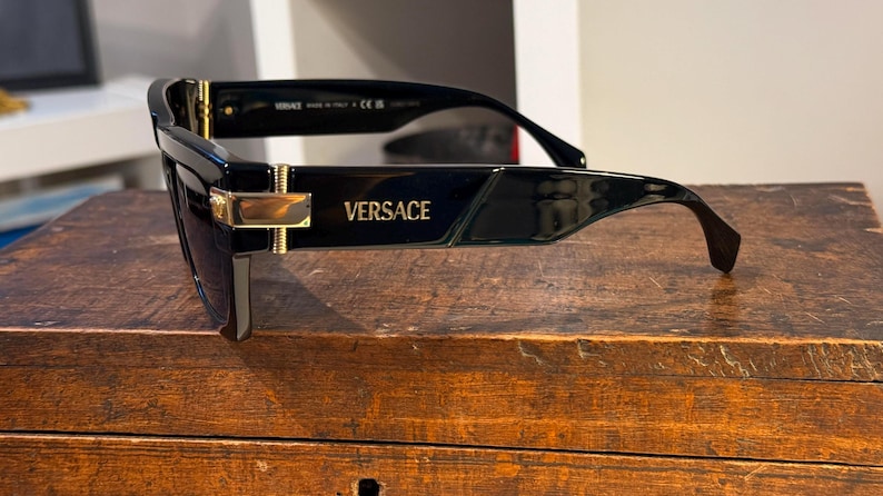 May include: Black Versace sunglasses with a rectangular frame and gold accents. The brand name is visible on the temple. The sunglasses are set on a wooden surface.