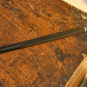 May include: A black mechanical pencil with silver detailing lies on a weathered wooden surface. The pencil features a textured grip and a metal tip. A small blue toy car is in the background.