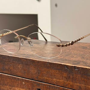 May include: Rimless eyeglasses with gold-colored frames and decorative accents. The temples feature a series of small, dark-colored embellishments. The lenses are oval-shaped and the glasses rest on a wooden surface.