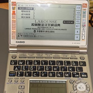 Puede incluir: Un diccionario electrónico Casio E-SF400 de color plateado con una pantalla abatible que muestra la interfaz del diccionario francés-chino Larousse. El dispositivo cuenta con un teclado QWERTY con varias teclas de función, un panel de navegación y una pequeña pantalla LCD secundaria debajo del área de visualización principal.
