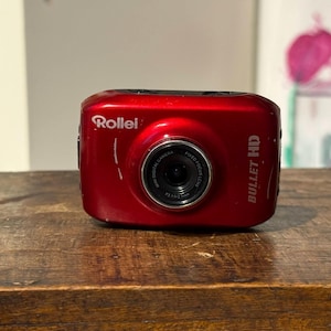 Puede incluir: Una cámara roja Rollei Bullet HD con lente negra, sobre una superficie de madera. La cámara tiene el logotipo de Rollei y "BULLET HD" impreso. El fondo está borroso, con obras de arte.