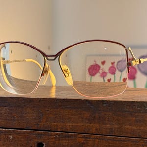 Peut inclure: Lunettes avec une monture dorée et un bord supérieur bordeaux. Les verres transparents reposent sur une surface en bois. Les branches des lunettes sont dorées. Design vintage.