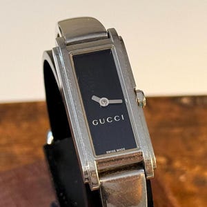 May include: A silver-toned Gucci wristwatch with a rectangular black watch face. The watch face displays the brand name "GUCCI" and "SWISS MADE". The watch has a silver band and a minimalist design.