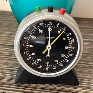 May include: A round analog Peter brand timer with a black face, white numerical markings, and a silver metallic frame. It features three small buttons on top in green, black, and red, with yellow hands and a black base. The text reads peter and Made in Germany.