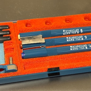 Könnte beinhalten: Eine blaue Kunststoffbox mit orangefarbenem Samtinterieur enthält mehrere Zeichenstifte und Zubehör. Die Stifte sind blau und mit "STAEDTLER MARS-730" beschriftet. Tintenpatronen und ein kleines blaues Werkzeug sind ebenfalls enthalten. Die Box ist geschlossen.