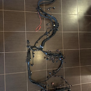 May include: A black automotive wiring harness with multiple connectors and wires. The harness is laid out on a brown tiled floor.