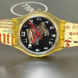 May include: A Swatch watch with a transparent yellow case and band. The watch face is black with white numerals and red hands. The band has printed text in red and black. The watch is a vintage style.