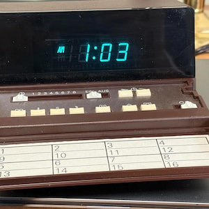 May include: A brown digital alarm clock with a black display screen showing the time 1:03 in green LED digits. The device features a control panel with beige buttons, a sliding switch, and a pull-out tray containing a grid of numbered squares.