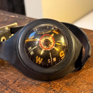 May include: A vintage black compass watch with a leather strap and a gold buckle. The compass face is black with white and yellow markings, including the cardinal directions. The watch is set on a wooden surface.