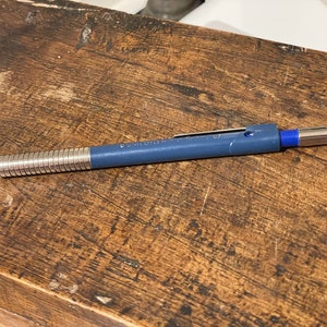May include: A blue mechanical pencil with a silver retractable tip and a blue eraser. The pencil is lying on a wooden surface.