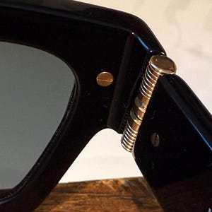May include: Close-up of black sunglasses with a gold hinge and screws. The lenses are a dark gray color. The sunglasses are likely for fashion or sun protection.