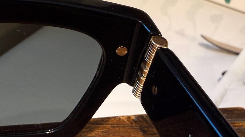 May include: Close-up of black sunglasses with a gold hinge and screws. The lenses are a dark gray color. The sunglasses are likely for fashion or sun protection.