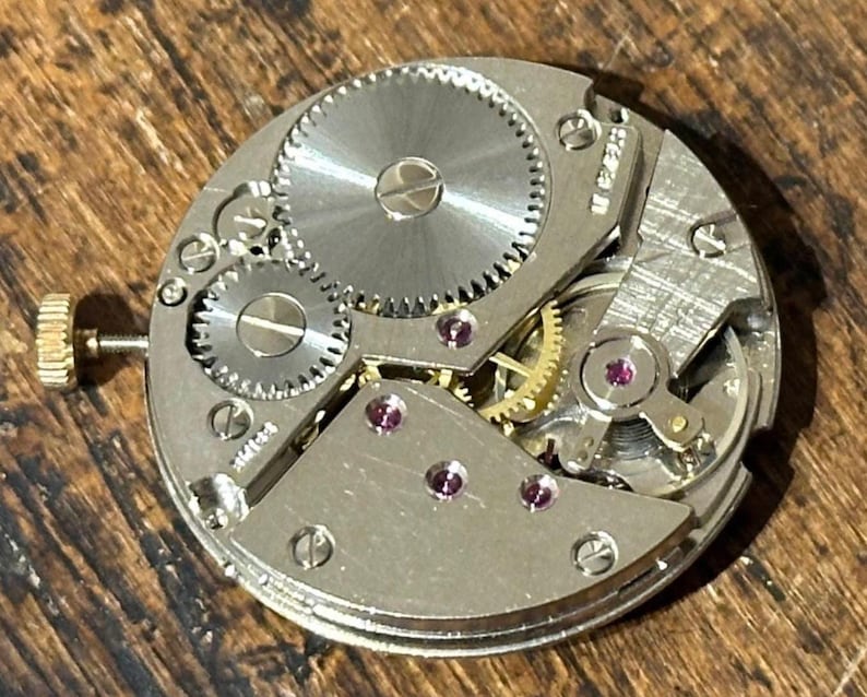 Pu&ograve; includere: Primo piano di un movimento di orologio vintage, con ingranaggi intricati, componenti in metallo argentato e piccole gemme di rubino. Il meccanismo dell'orologio &egrave; posto su una superficie di legno, evidenziando la lavorazione artigianale.