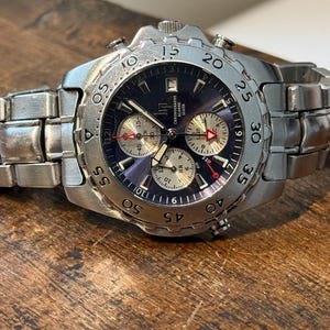 Puede incluir: Un reloj cronógrafo de acero inoxidable en tono plateado con esfera azul oscuro, tres subesferas y un bisel giratorio con números grabados. El reloj incluye una correa de eslabones metálicos, manecillas luminosas y una ventana de fecha.