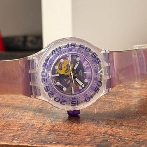 Könnte beinhalten: Eine transparente lila Armbanduhr mit einem klaren Zifferblatt und einer lila Lünette. Die Uhr hat ein lila Armband und einen sichtbaren Innenmechanismus mit goldenen und schwarzen Komponenten. Die Uhr liegt auf einer Holzoberfläche.