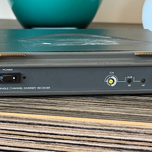 Peut inclure: Un récepteur de diversité monocanal JTS US-8001D UHF PLL gris avec deux petites antennes horizontales, un interrupteur d'alimentation noir, un cadran de sélection de canal, des voyants A/B et un bouton de volume noir sur le panneau avant.