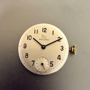 K&ouml;nnte beinhalten: Ein Vintage-Zifferblatt mit cremefarbenem Zifferblatt, schwarzen Zeigern und arabischen Ziffern. Das Zifferblatt hat eine kleine Sekundensubskala und eine goldfarbene Krone. Der Markenname "RECORD" ist auf dem Zifferblatt aufgedruckt. "SWISS MADE" ist unten aufgedruckt.