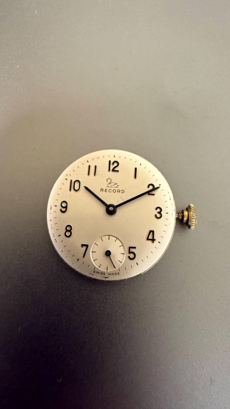 K&ouml;nnte beinhalten: Ein Vintage-Zifferblatt mit cremefarbenem Zifferblatt, schwarzen Zeigern und arabischen Ziffern. Das Zifferblatt hat eine kleine Sekundensubskala und eine goldfarbene Krone. Der Markenname "RECORD" ist auf dem Zifferblatt aufgedruckt. "SWISS MADE" ist unten aufgedruckt.