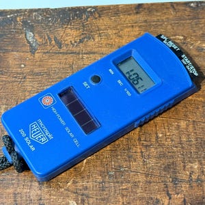 Puede incluir: Un cronómetro azul Heuer Microsplit 200 Solar con un cordón negro. La pantalla digital muestra un tiempo de 1:05.11. El cronómetro tiene una célula solar y varios botones para configurar y cronometrar.